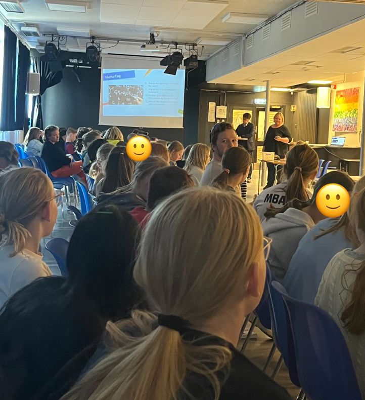en gruppe mennesker som sitter i et rom med en person som står foran en skjerm